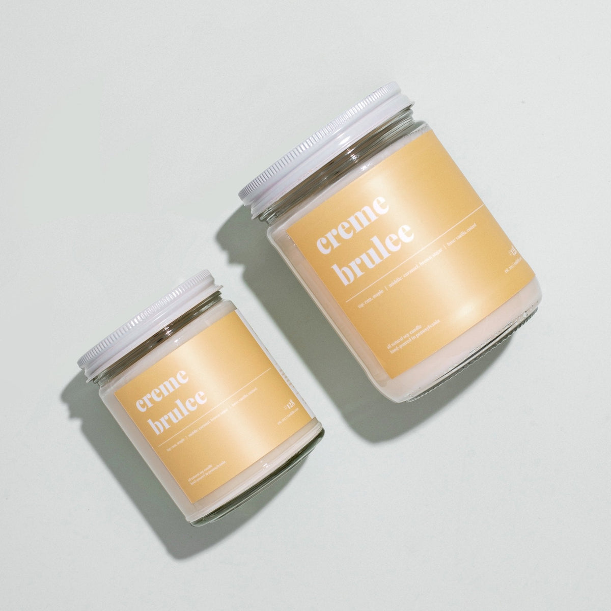 Overhead view of petite and standard creme brulee soy candles with white lids on a pale grey surface