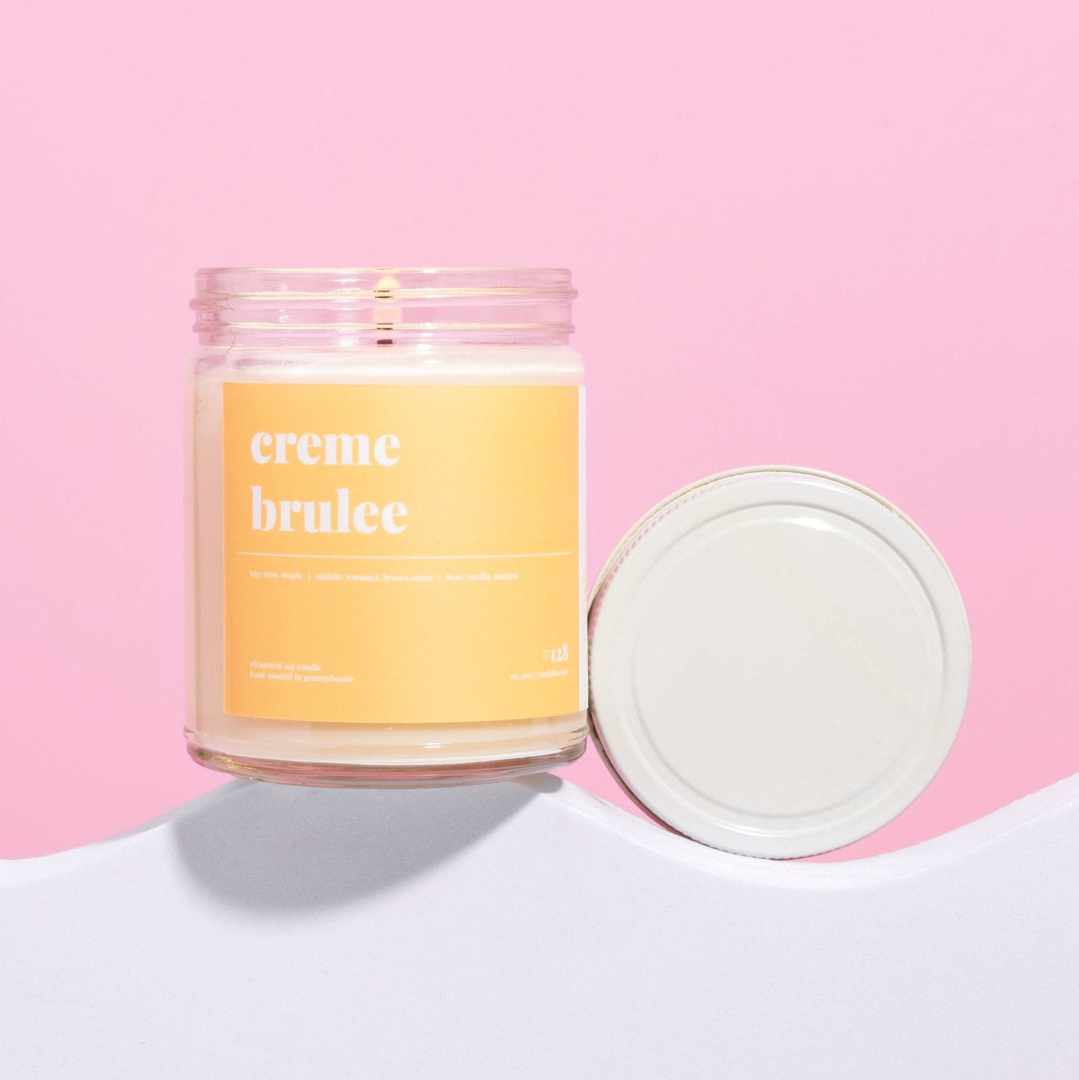 Creme brulee soy candle burning in a clear jar with a soft yellow label and white lid leaning beside it on pink