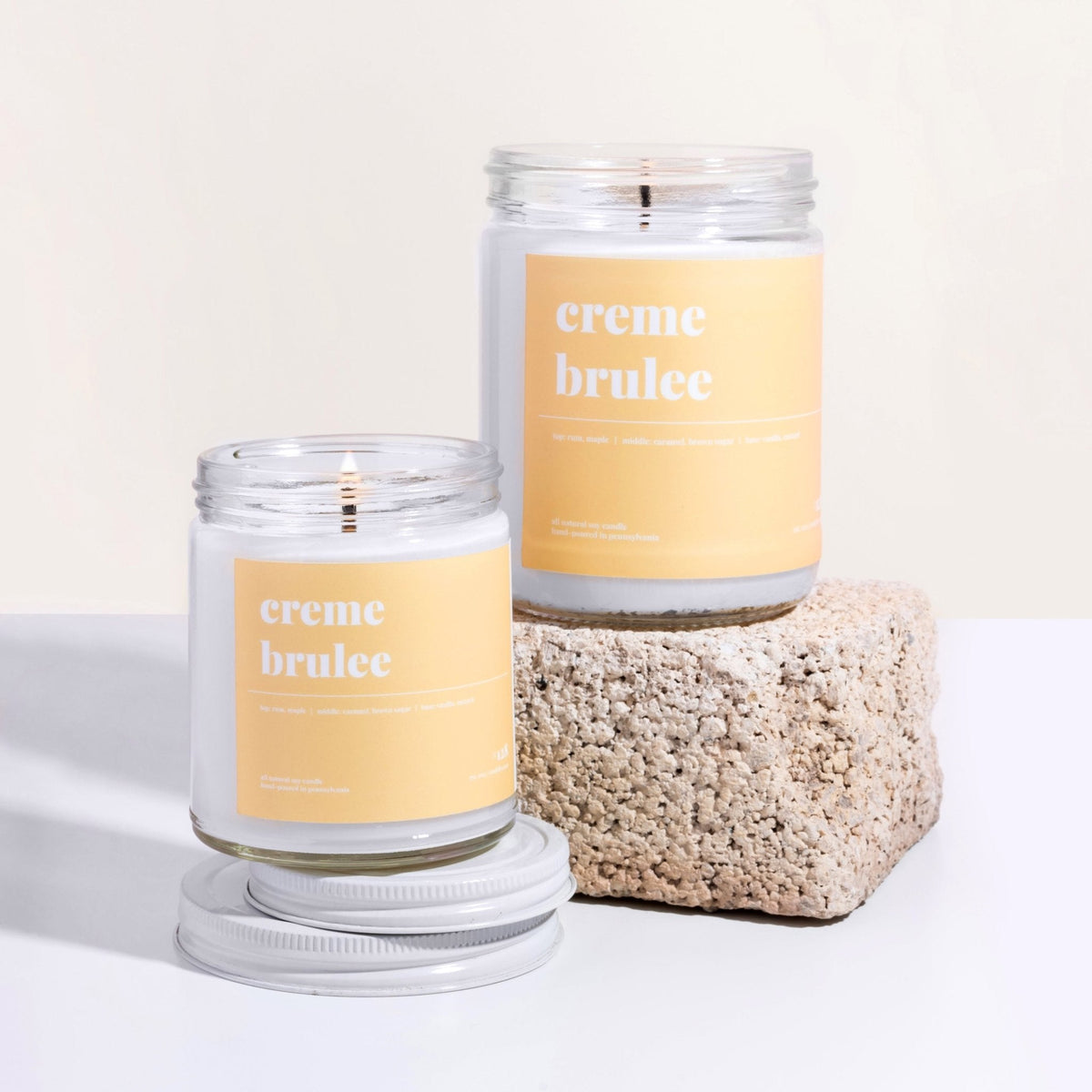 Two creme brulee soy candles with yellow labels on a textured stone riser, the smaller jar lit and open