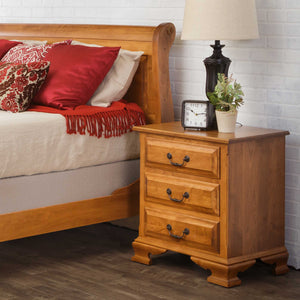 Three-drawer oak nightstand with black bail pulls and ogee bracket feet beside a sleigh bed with red pillows