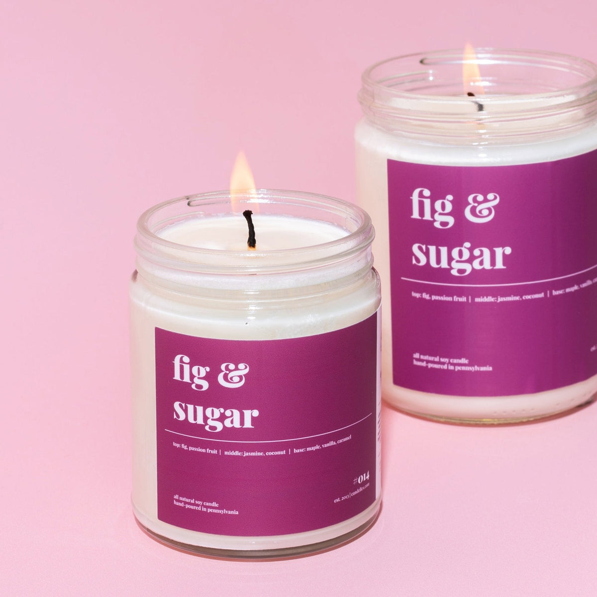 Two fig and sugar soy candles burning in clear jars with deep magenta labels on a soft pink background
