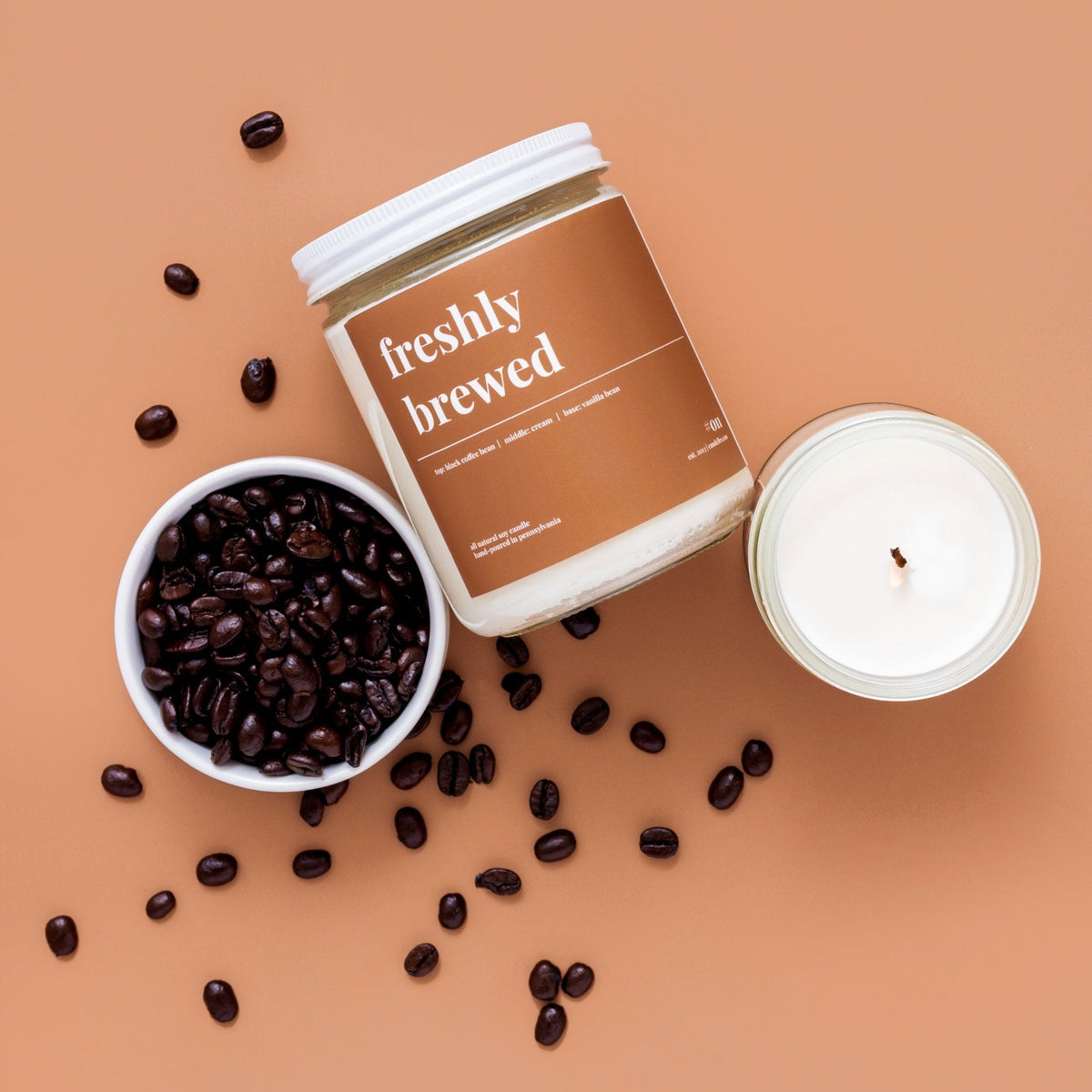 Freshly brewed soy candle beside an open jar and a bowl of dark roasted coffee beans scattered on terracotta