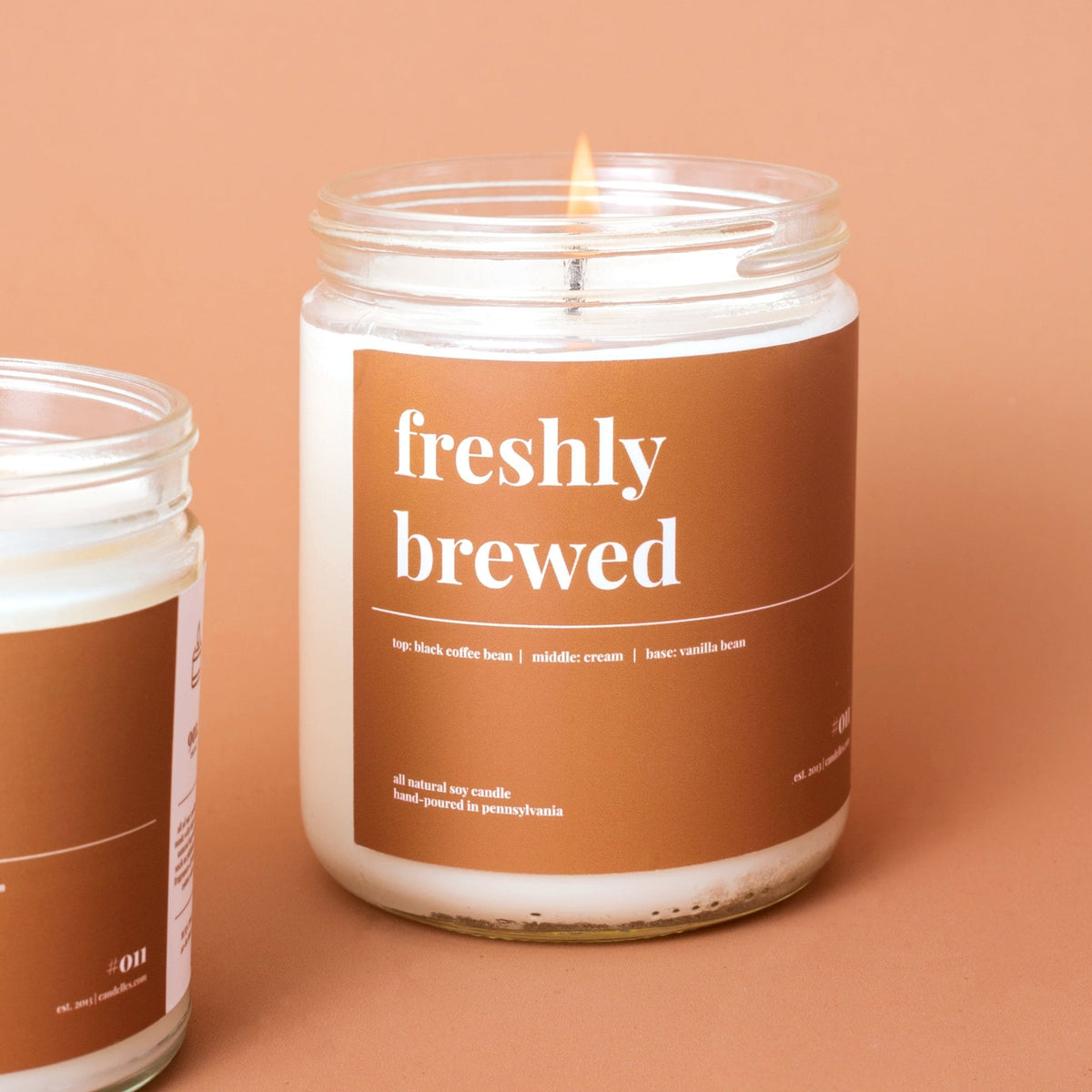 Close-up of a lit freshly brewed coffee soy candle with brown label next to a second jar on terracotta