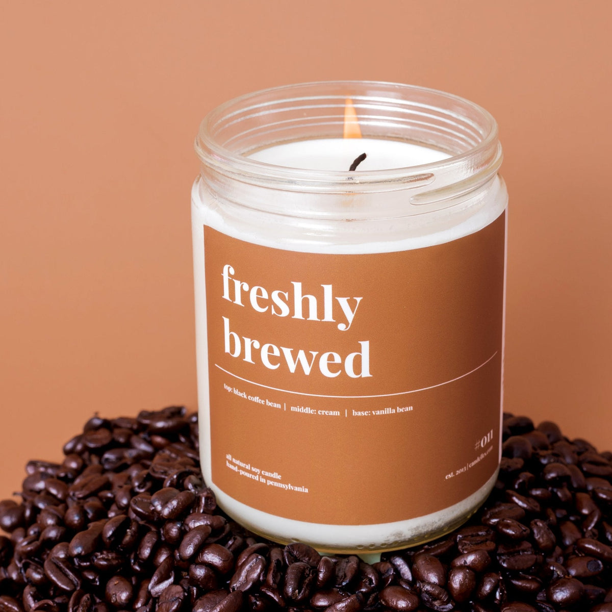 Lit freshly brewed soy candle sitting on a mound of dark roasted coffee beans against a warm terracotta background