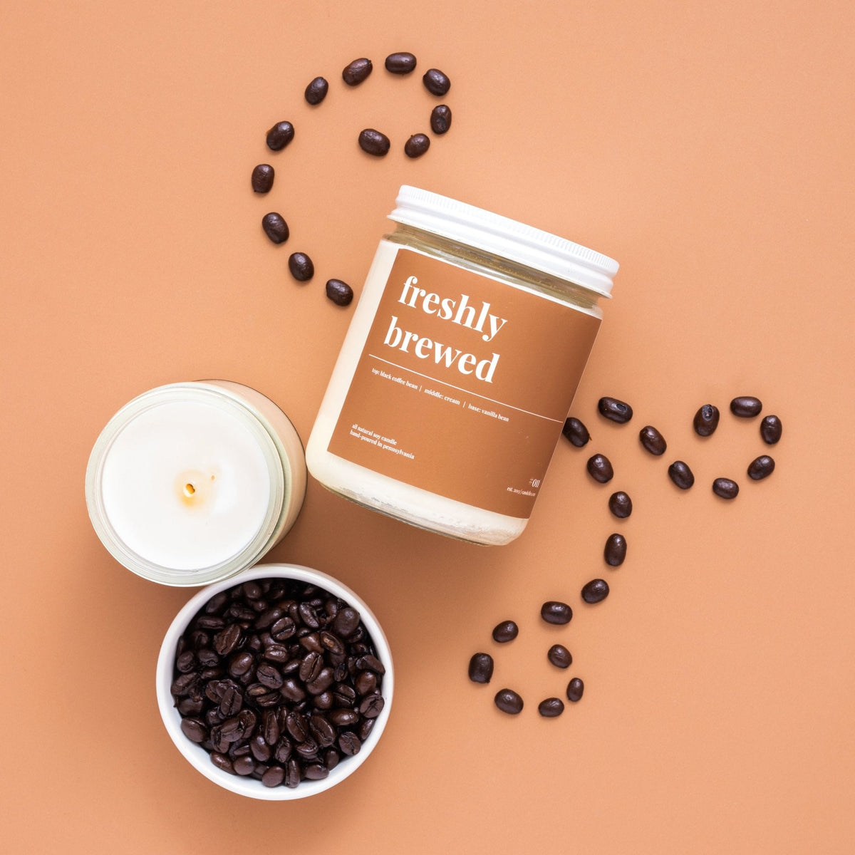Overhead freshly brewed candle with lid, a lit open jar and a bowl of coffee beans swirled across terracotta