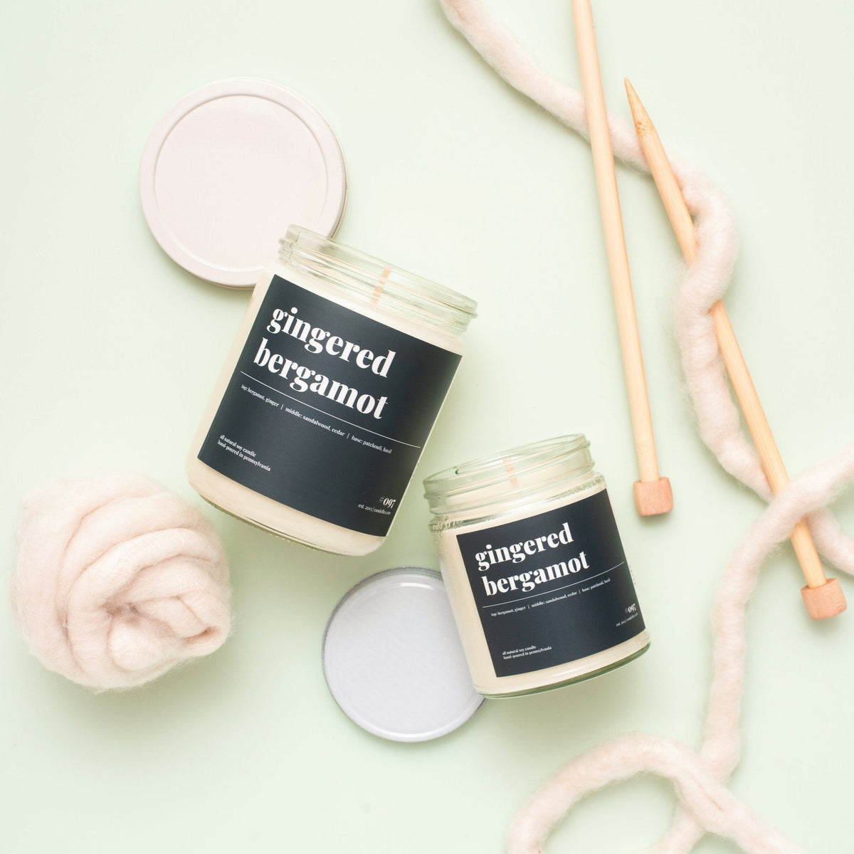 Two gingered bergamot soy candles with black labels and open lids beside wool roving and knitting needles on mint