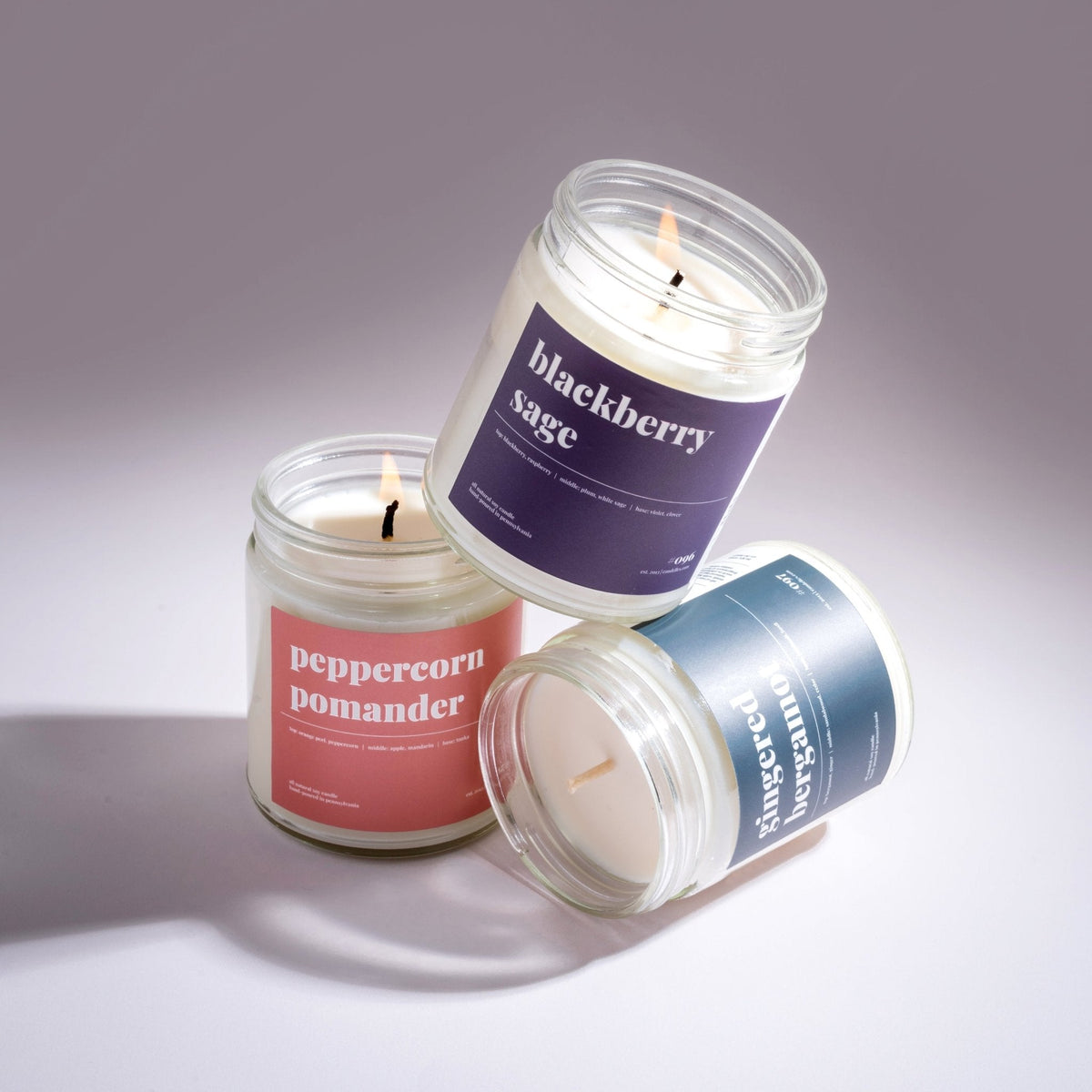 Three lit soy candles stacked at angles — peppercorn pomander, blackberry sage and gingered bergamot — on grey