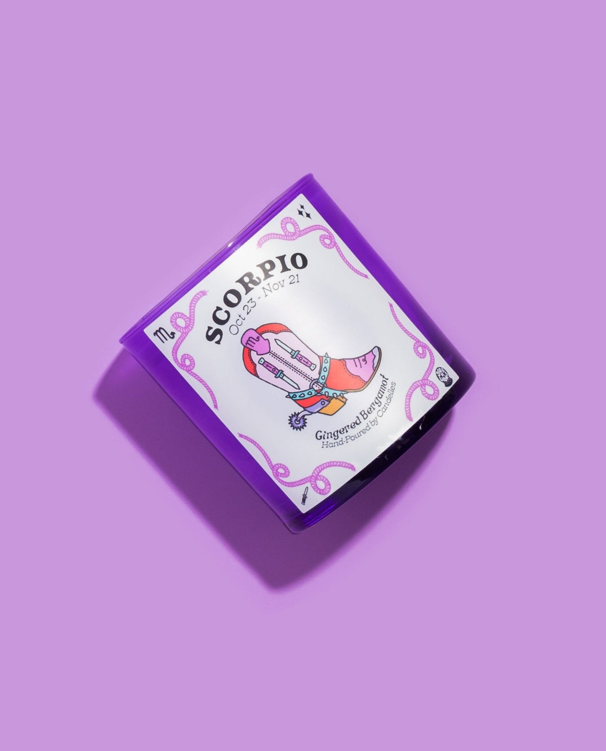 Scorpio gingered bergamot zodiac candle tilted on lilac, showing the illustrated cowboy boot and date label