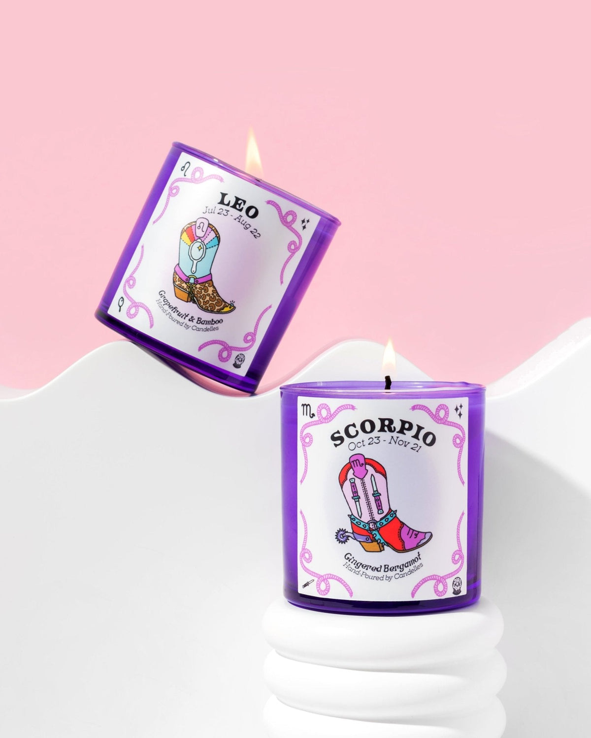 Leo and Scorpio zodiac candles in purple glass tumblers burning on white wave risers against a pink backdrop