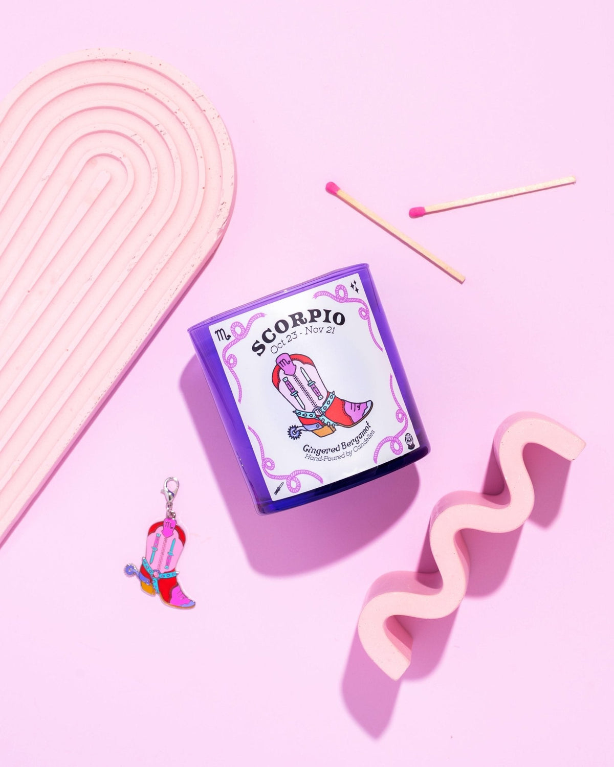 Gingered bergamot Scorpio zodiac candle in a purple glass tumbler with matches, boot charm and pink props on pink