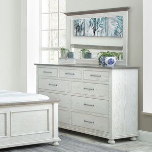 Whitewashed nine drawer dresser with gray top and wide mirror styled with plants beside a matching bed