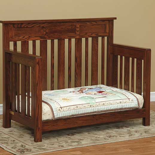 Oak crib in day bed configuration with open front, slat back and a printed child's quilt