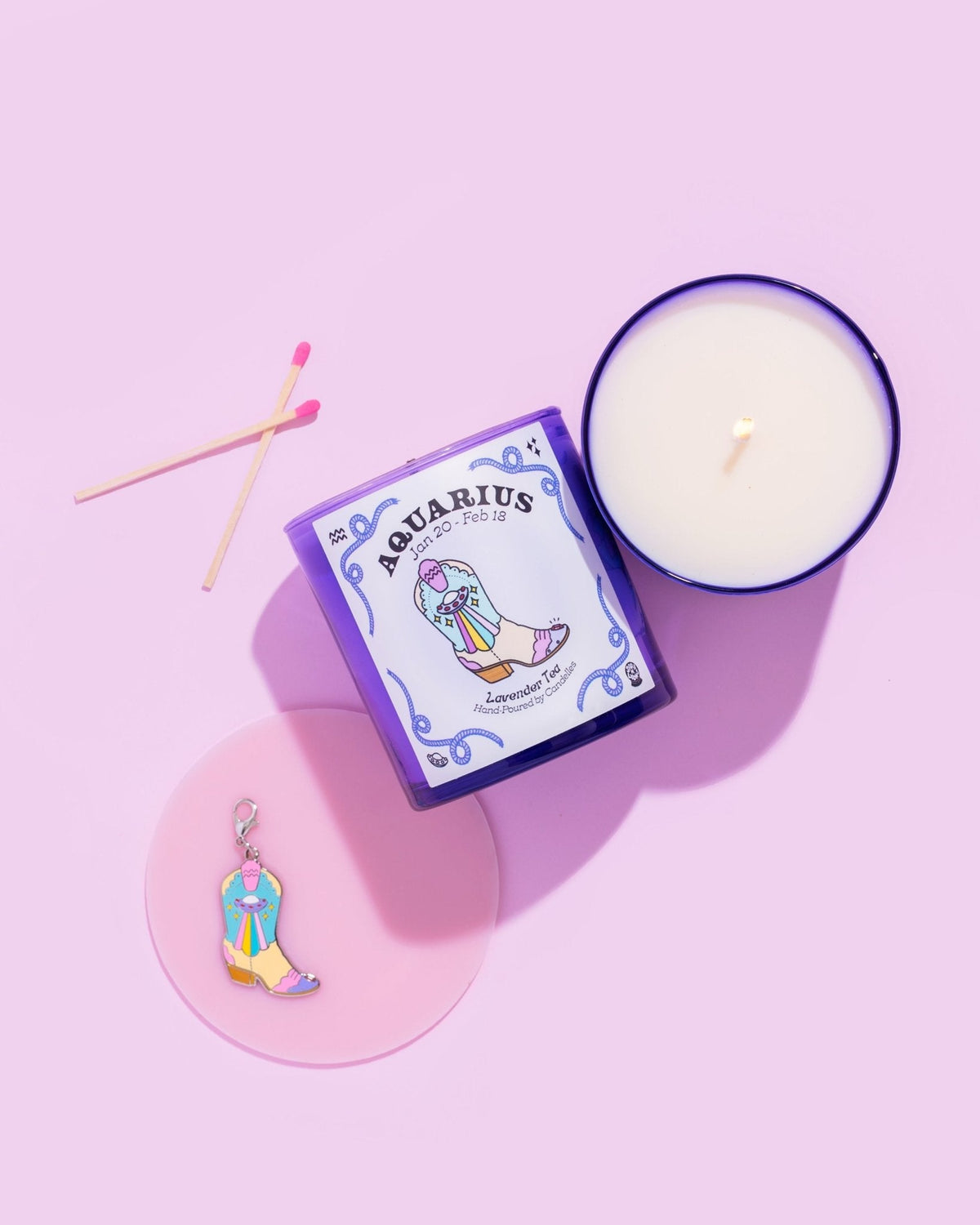Lavender tea Aquarius zodiac candle with matches, a boot charm and a lit purple glass jar on a pink background