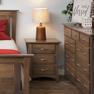 Curved leg nightstand in gray brown maple with three drawers and a lamp beside a bed and dresser