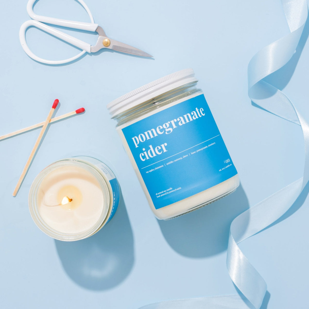 Flat lay of a lidded pomegranate cider candle with a lit open jar, scissors, matches and blue satin ribbon