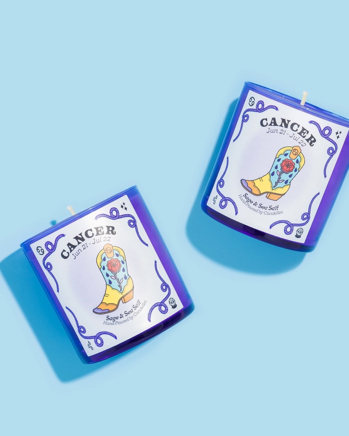 Two sage and sea salt Cancer zodiac candles in blue glass tumblers with illustrated boot labels on light blue