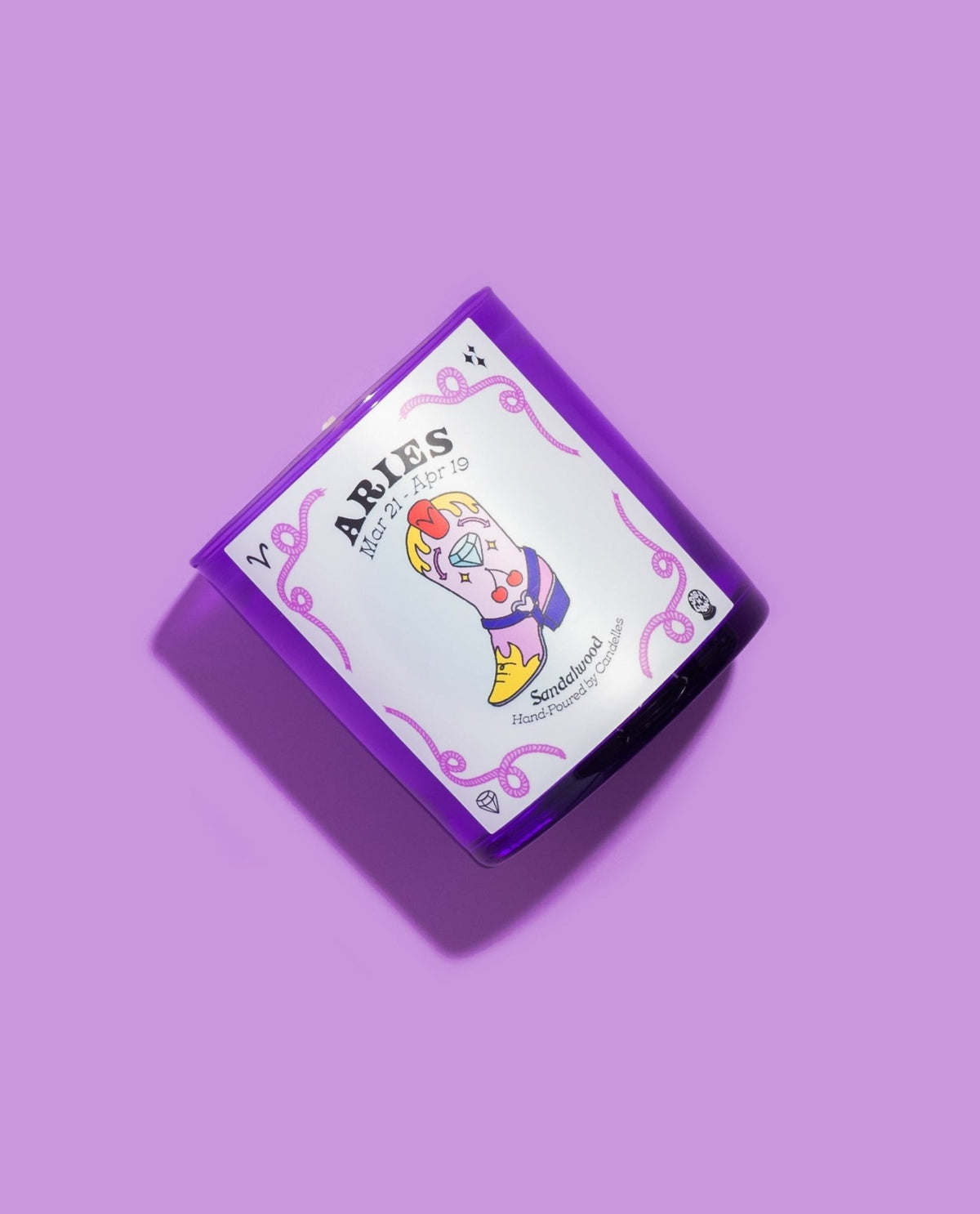 Sandalwood Aries zodiac candle angled on a lilac backdrop, showing the pink illustrated cowboy boot label