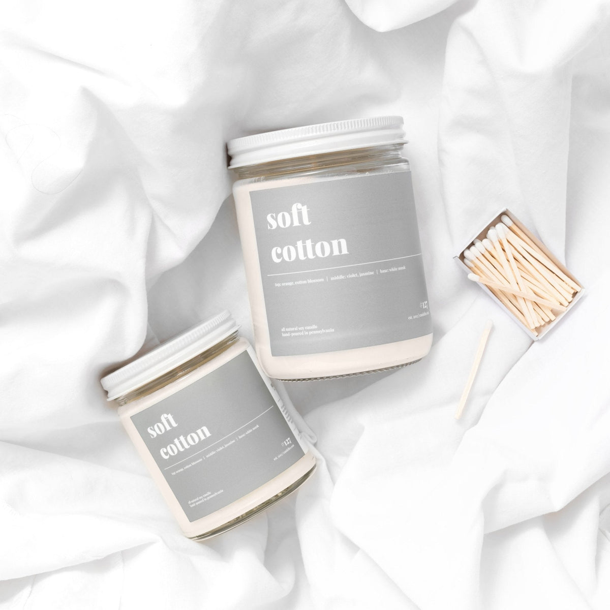 Petite and standard soft cotton candles with white lids resting on rumpled white bedding beside a matchbox