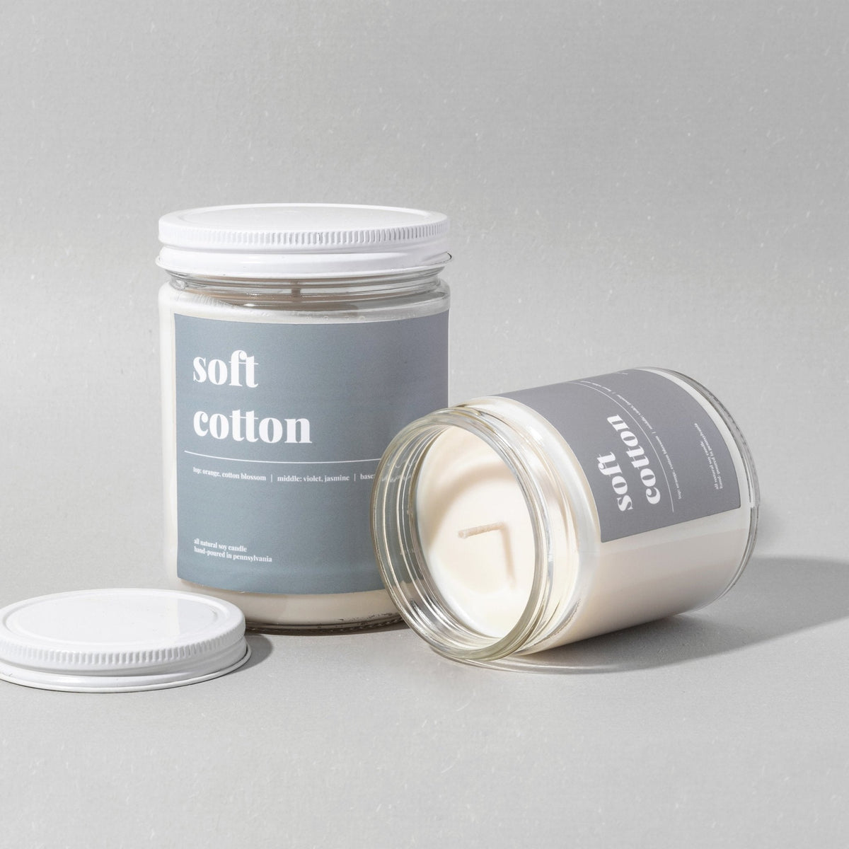 Standard soft cotton soy candle upright beside a jar tipped on its side showing the wick, with lids on grey
