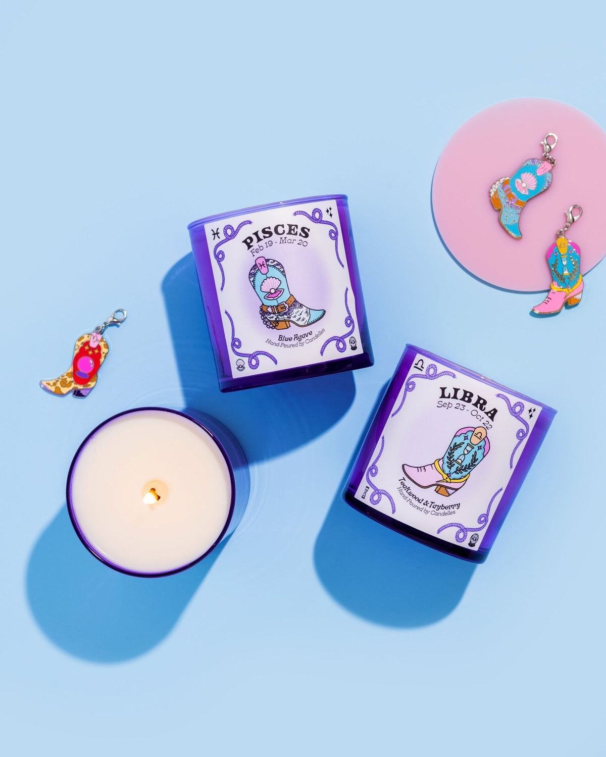 Pisces and Libra purple zodiac candles laid flat with cowboy boot charms and one lit jar on a blue backdrop