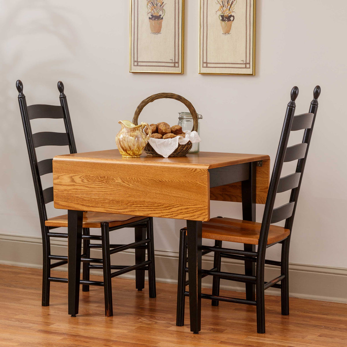 A small wooden dining table with a drop-leaf design and two black ladder-back chairs with wood seats, shown in a staged dining room setting with framed wall art and a wood floor.