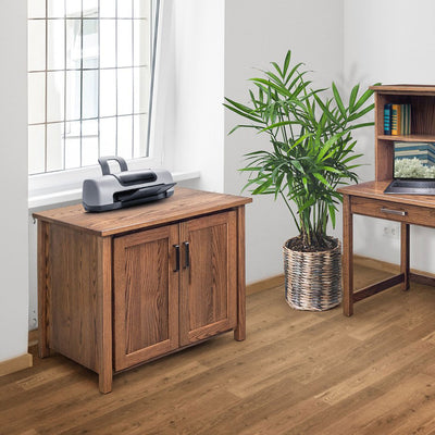 Oak printer cabinet with two paneled doors and dark bar pulls, holding a printer beside a sunny home office window