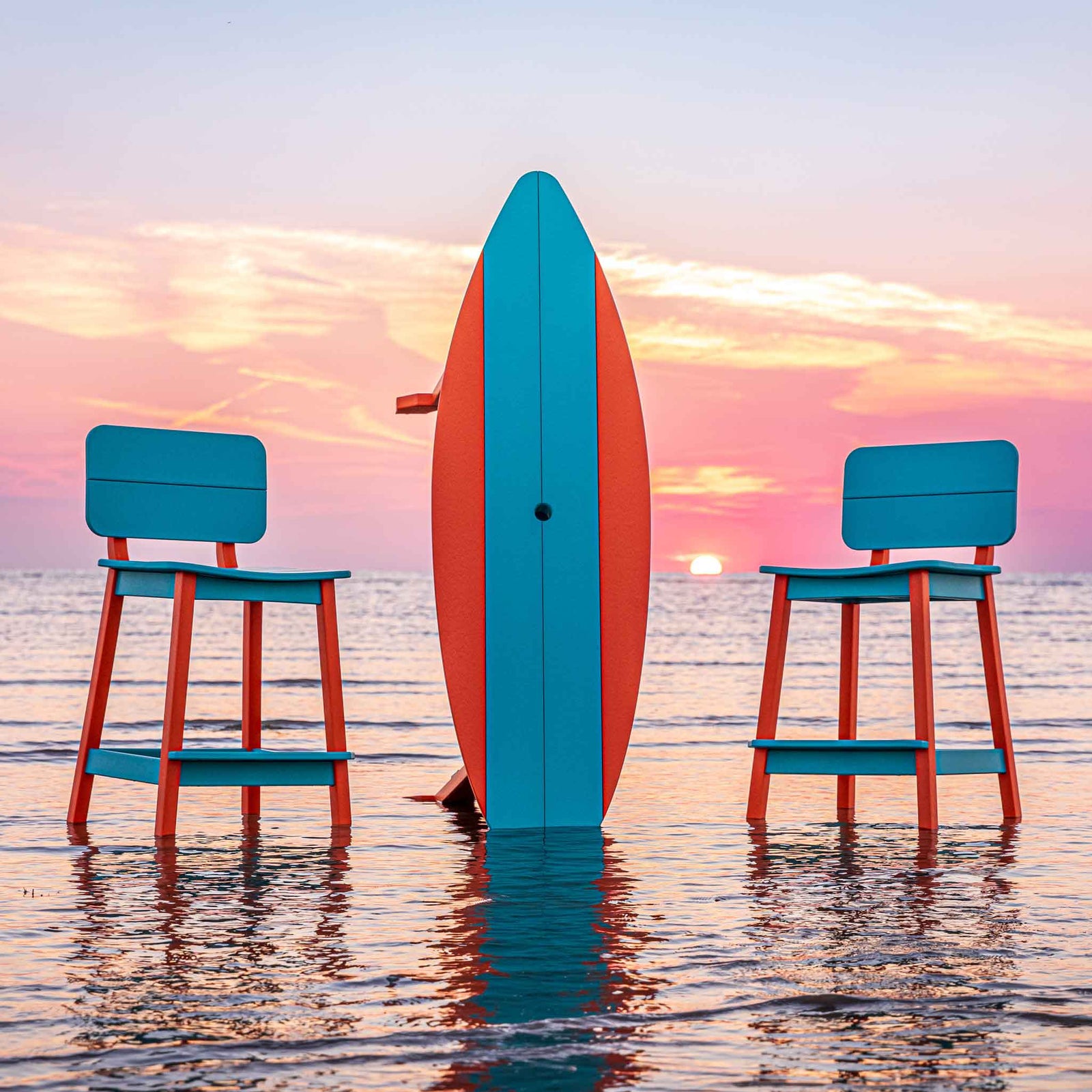 Surfboard table balanced on its tail with two turquoise bar stools at either side at sunset