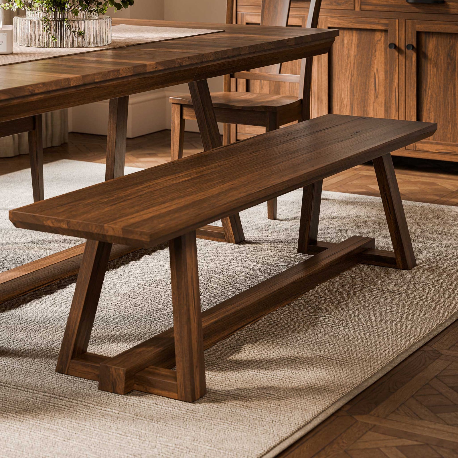 Long walnut bench with trestle legs and stretcher beside a matching dining table on a herringbone wood floor