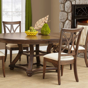 X back dining side chairs in weathered oak with cream upholstered seats around a round pedestal table by a stone fireplace