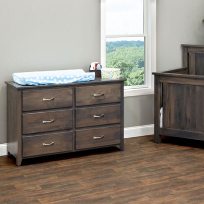 Gray six-drawer dresser topped with a blue changing pad beside a matching crib in a nursery with a bright window