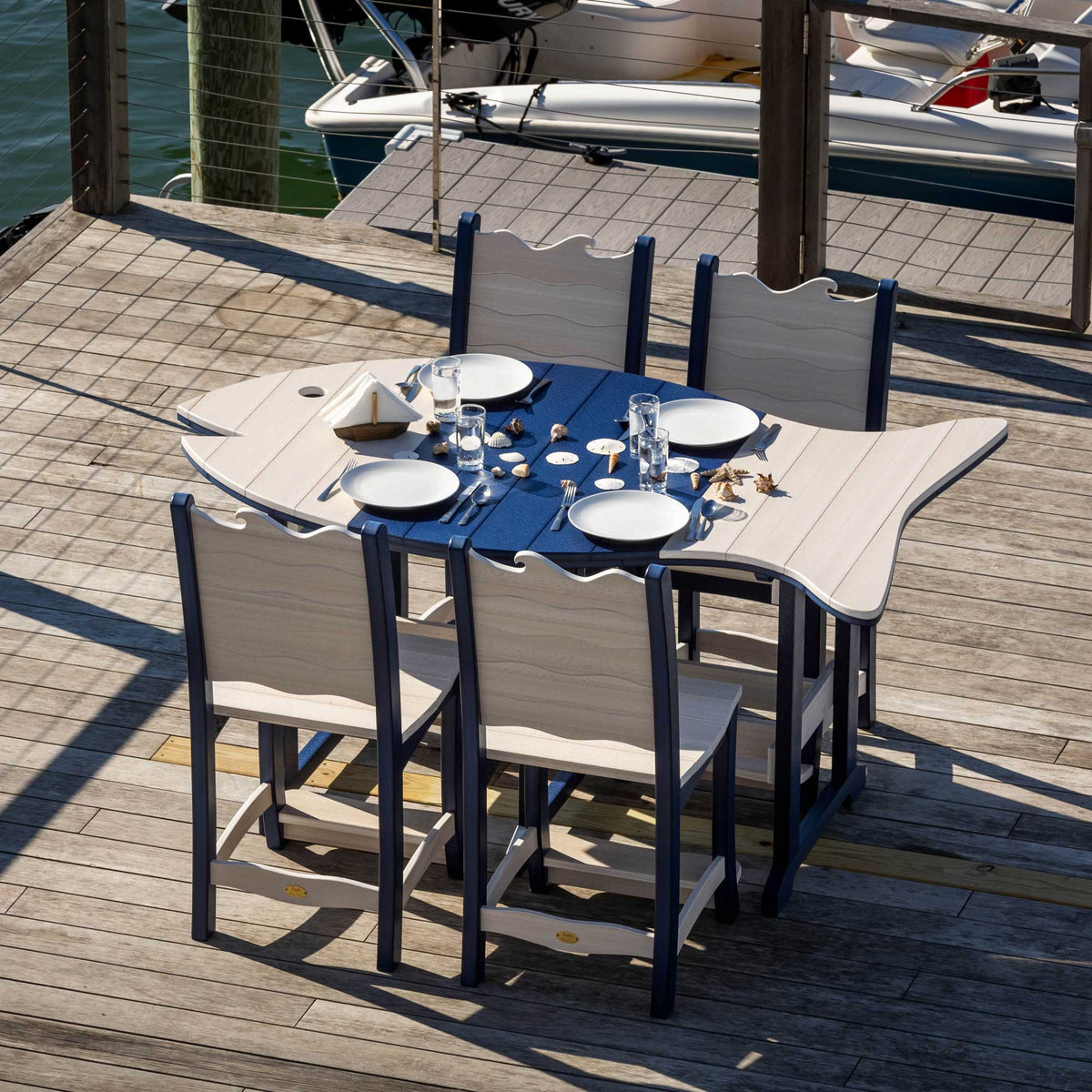 Large fish shaped poly dining table in navy and driftwood set with white plates and four matching chairs on a dock