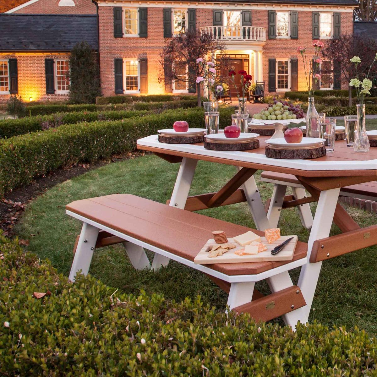 Cedar and white poly picnic table with benches set with apples and grapes on a lawn before a brick manor