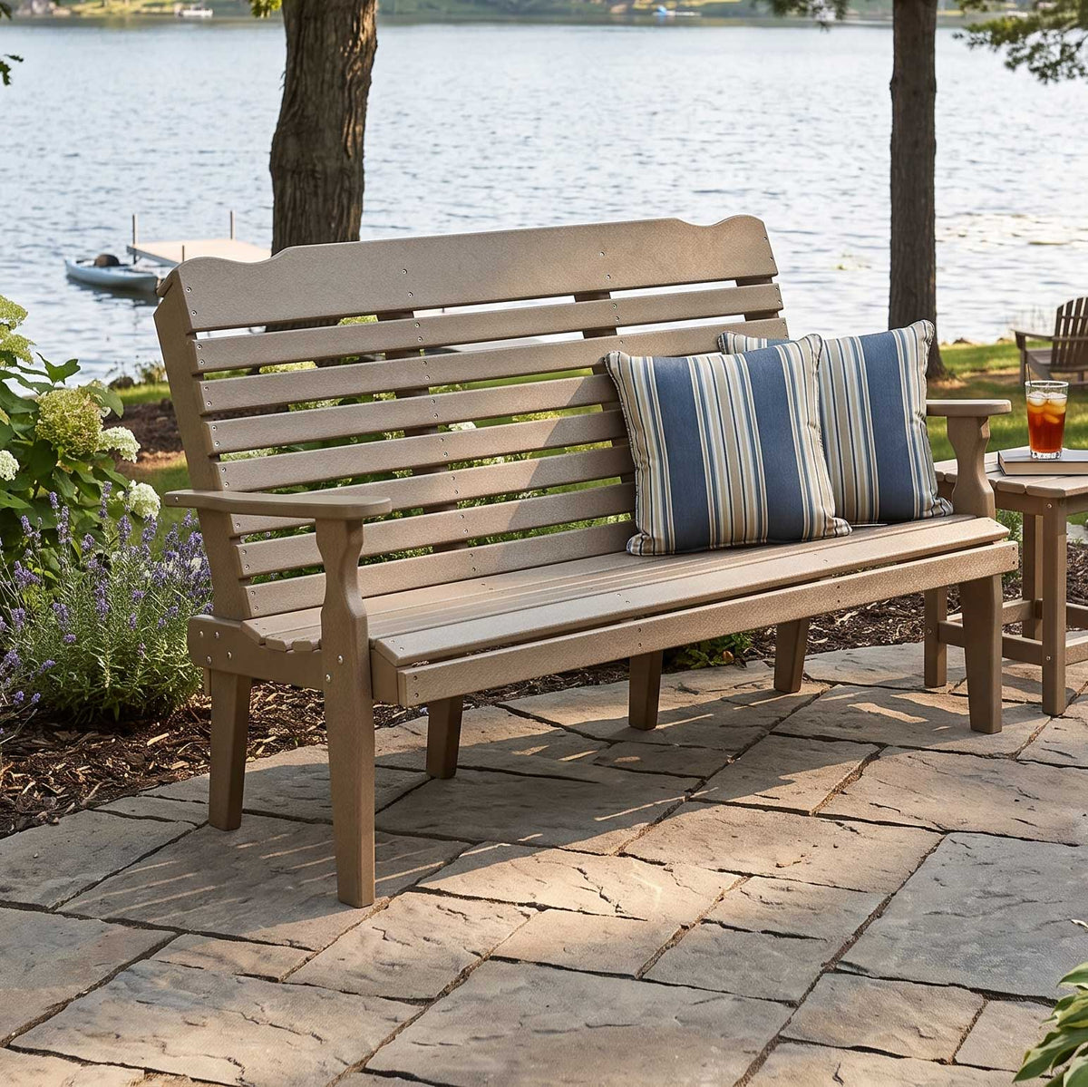Weatherwood poly 5 foot patio bench with slatted curved back and blue striped pillows on a flagstone patio overlooking a lake