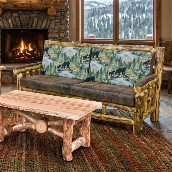 A rustic lodge-style living room featuring a stone fireplace, log furniture, and large windows with mountain views. The room includes a log chair, log sofa with patterned cushions, a log coffee table, a side table, and a woven area rug.