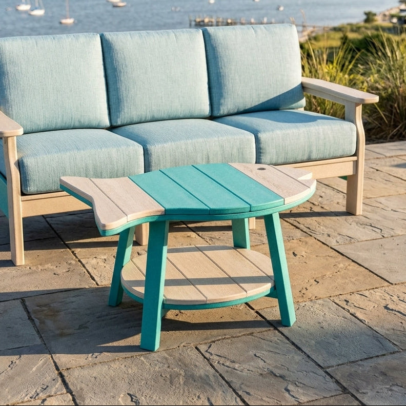 Outdoor patio furniture set with a three-seat cushioned sofa and a matching coffee table, shown on a stone deck overlooking a coastal bay with boats and shoreline.