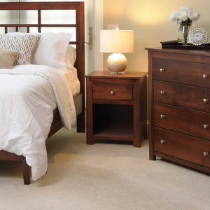 Cherry nightstand with one drawer and open shelf holding a lamp, beside a slat headboard bed and chest