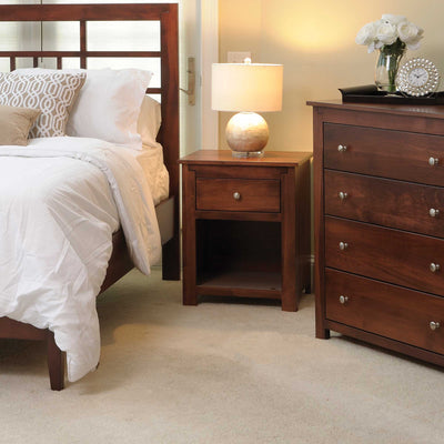 Cherry nightstand with one drawer and open shelf holding a lamp, beside a slat headboard bed and chest