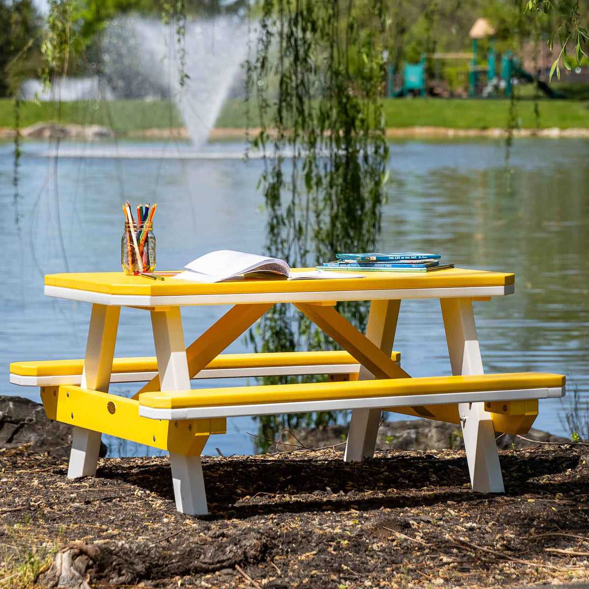 Bright yellow childrens picnic table with white A frame legs and attached benches at the edge of a lake