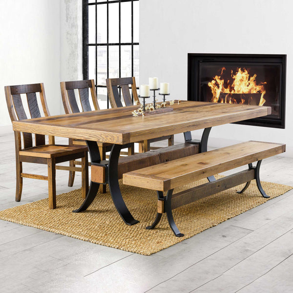 Reclaimed oak trestle farm table with black arched legs, matching bench and dark slat back chairs by a modern fireplace