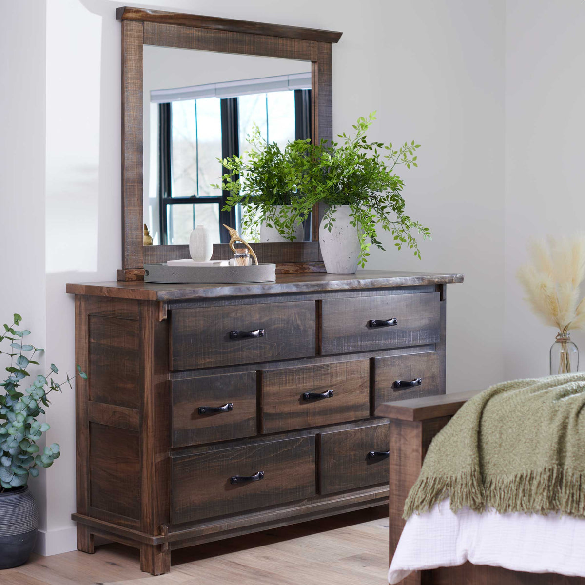 Rustic dresser with a large plank framed mirror reflecting a window, styled with a leafy vase in a white bedroom
