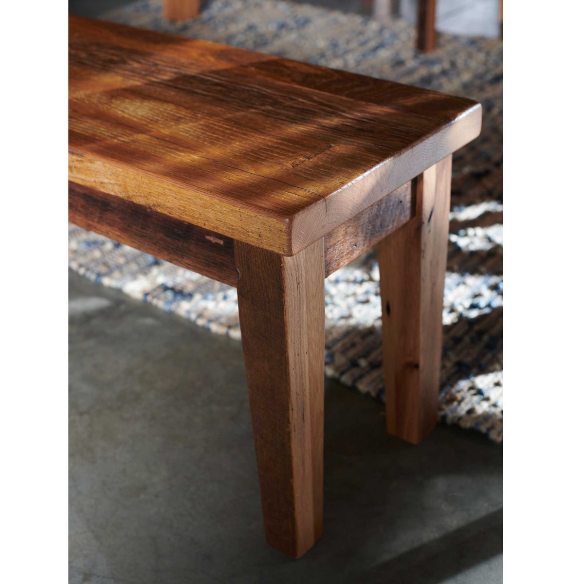 Close view of a barnwood bench corner showing saw marked plank seat and a tapered leg on a woven rug