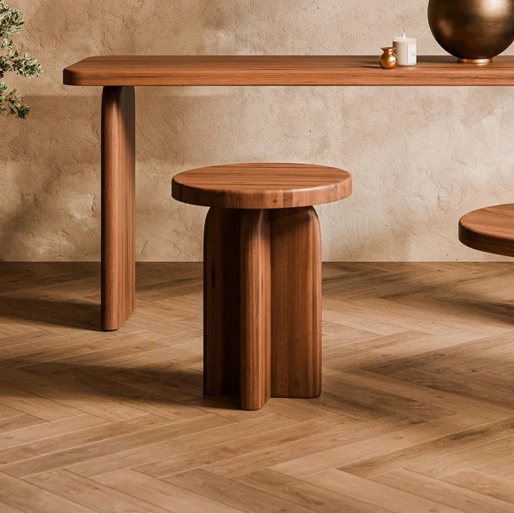 A warm, minimalist interior scene featuring a medium brown wooden console table, a matching round wooden stool, and an oval wooden coffee table with a sculptural block base. A white drum lamp with a metallic bronze base sits on the console table.