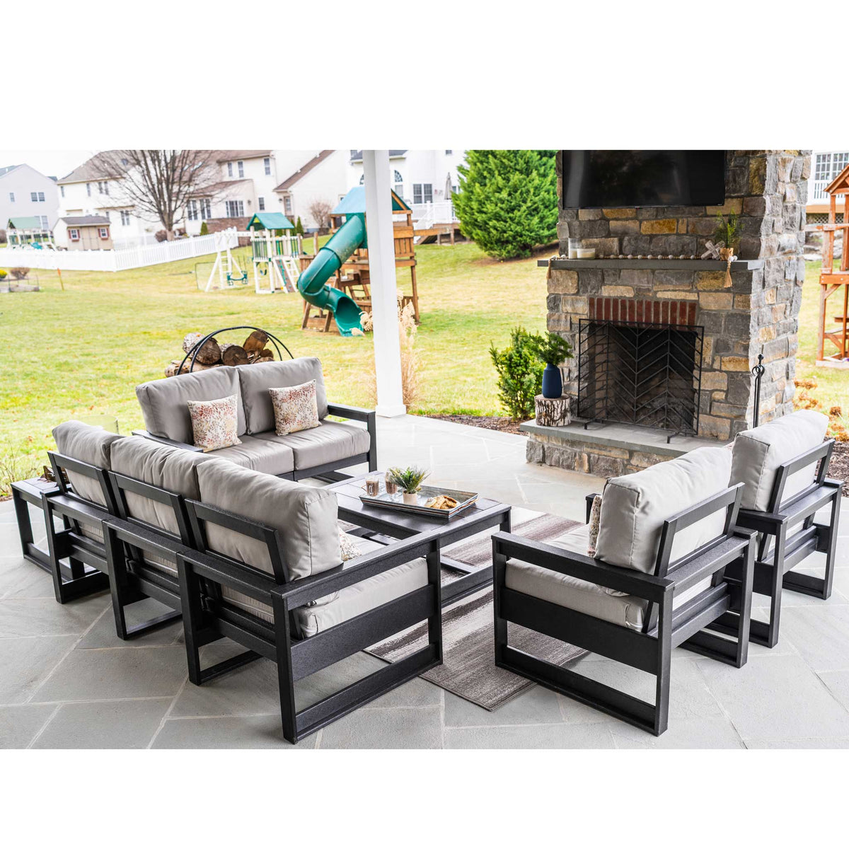 Black poly patio seating group with gray cushions on a stone patio beside a stone outdoor fireplace