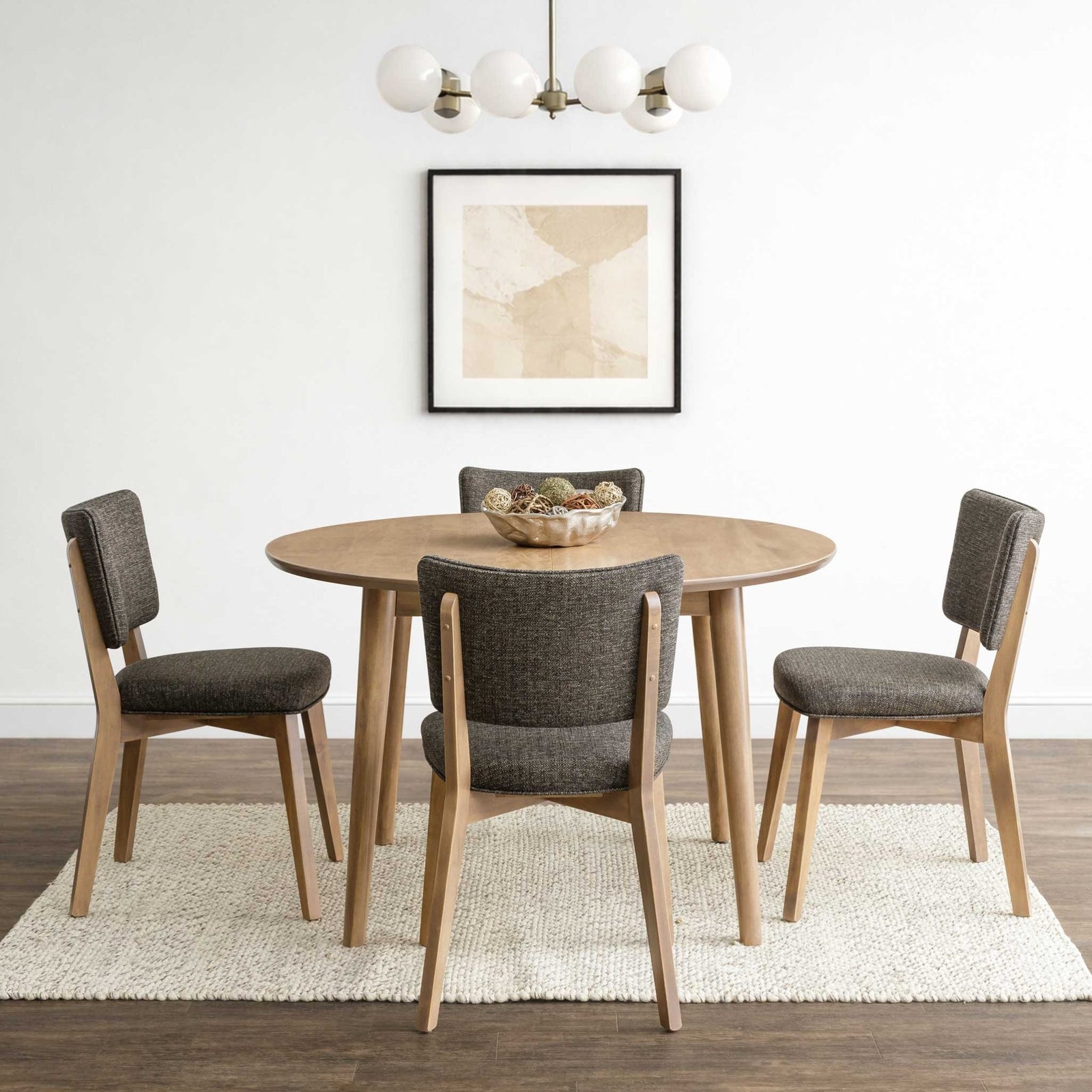 Round light maple dining table with four charcoal upholstered chairs on a woven rug under a globe chandelier