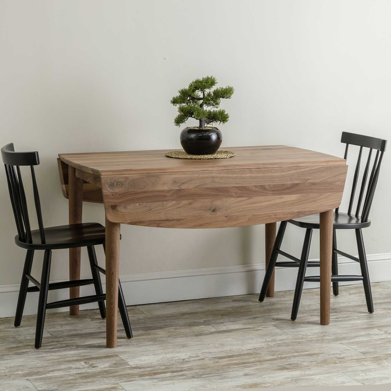 Walnut drop leaf table with one leaf down against a wall, paired with two black spindle back side chairs