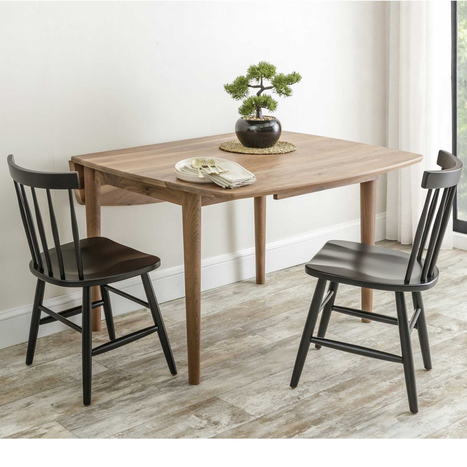 Walnut drop leaf table with one leaf down against a wall, paired with two black spindle back side chairs