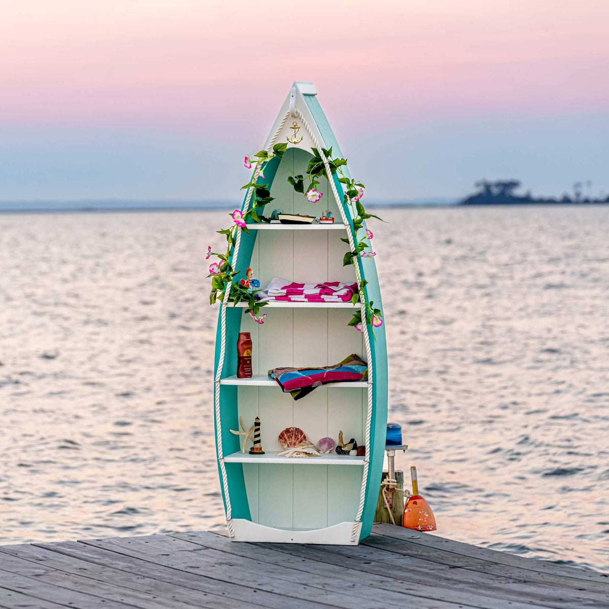 Teal and white boat bookcase on a dock at sunset, filled with beach towels, shells and nautical decor