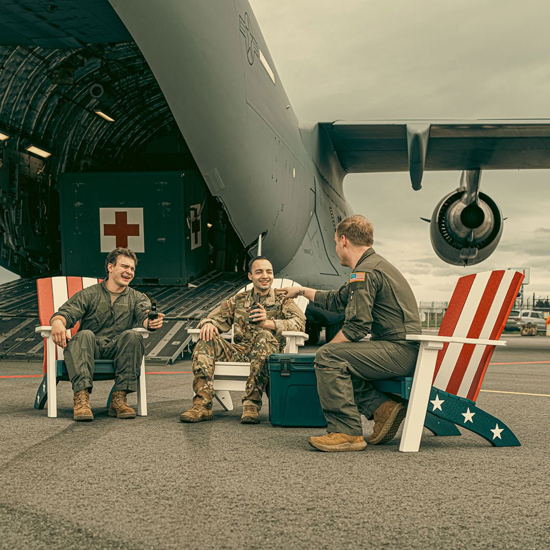 Three service members relaxing in red, white and blue poly Adirondack chairs beside a military cargo plane
