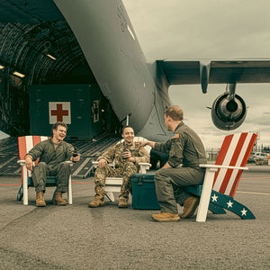 Three service members relaxing in red, white and blue poly Adirondack chairs beside a military cargo plane