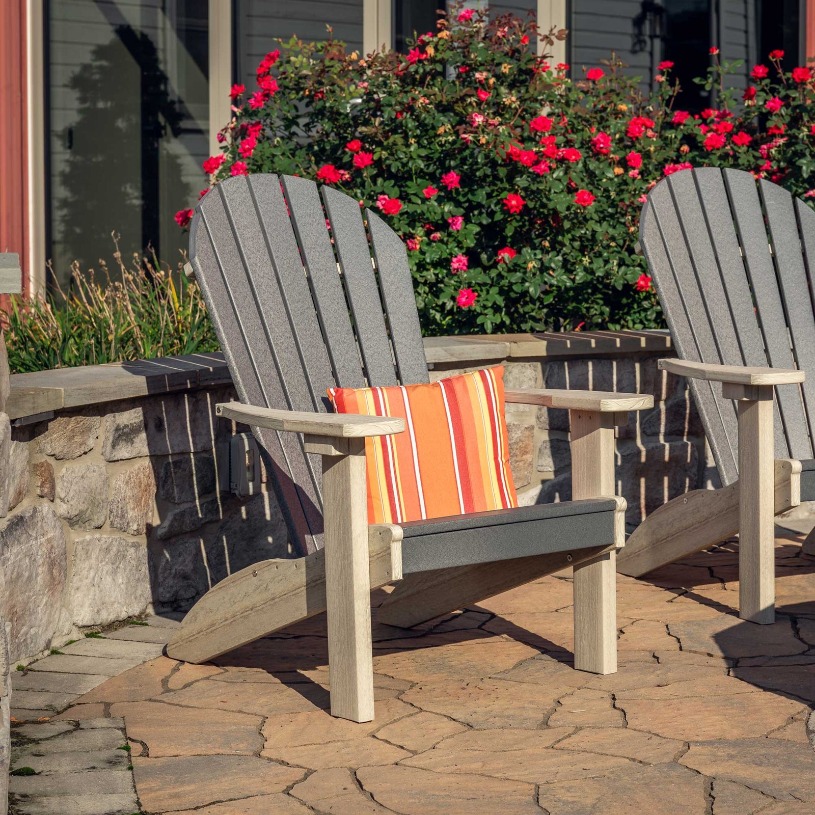 Gray and driftwood poly Adirondack chair with a striped orange pillow on a flagstone patio beside blooming roses